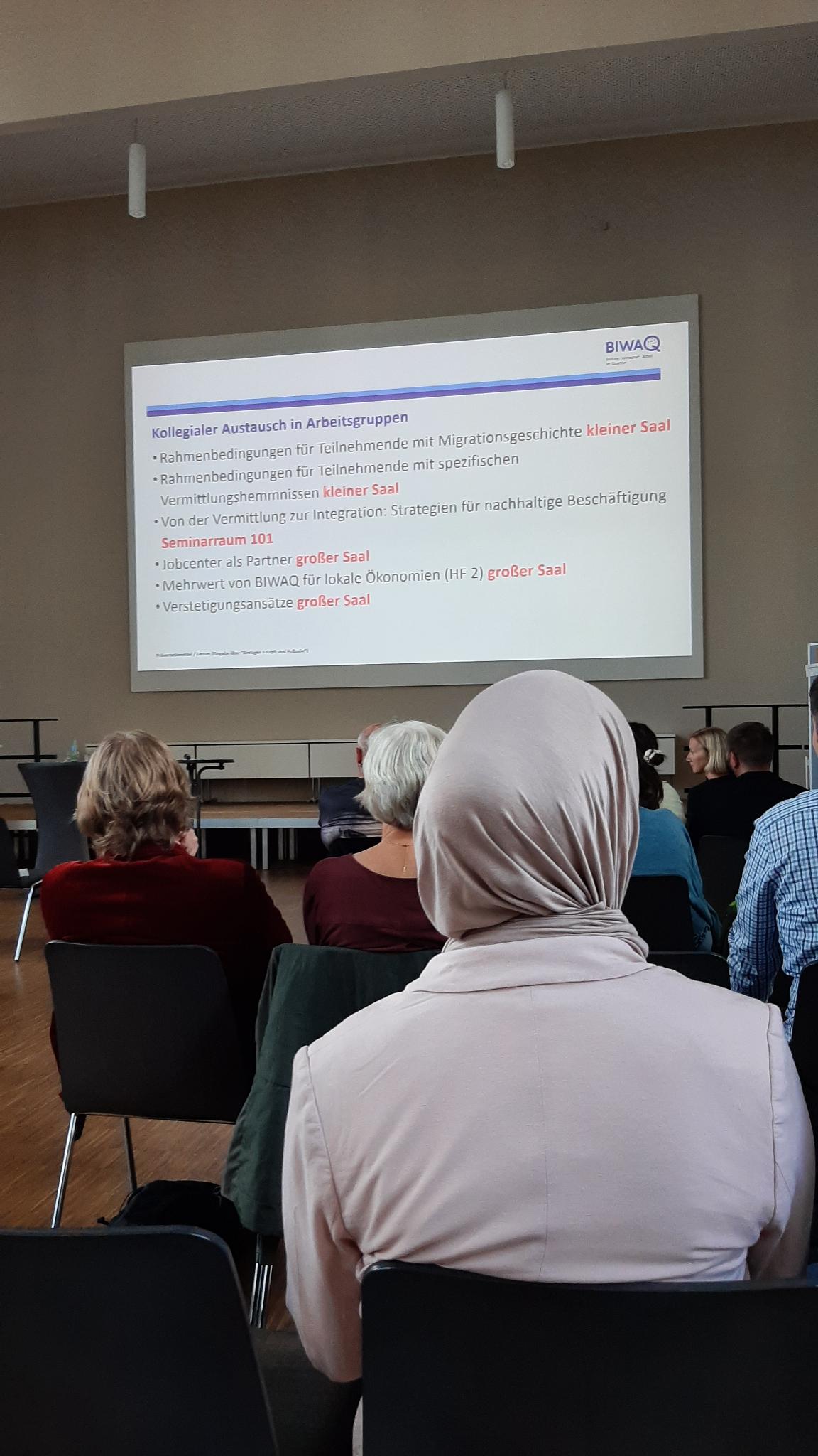 Menschen sitzen in einem Saal und sehen eine Leinwandpräsentation